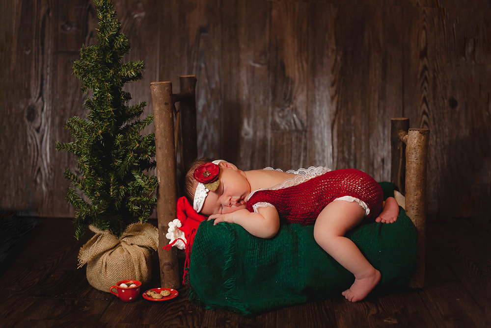 Newborn Baby girl in a frilly red onesie sleeps peacefully on a wooden frame bed with green sheets. Next to the bed is a tiny Christmas tree, hot cocoa in a tiny cup, and a tiny plate of cookies